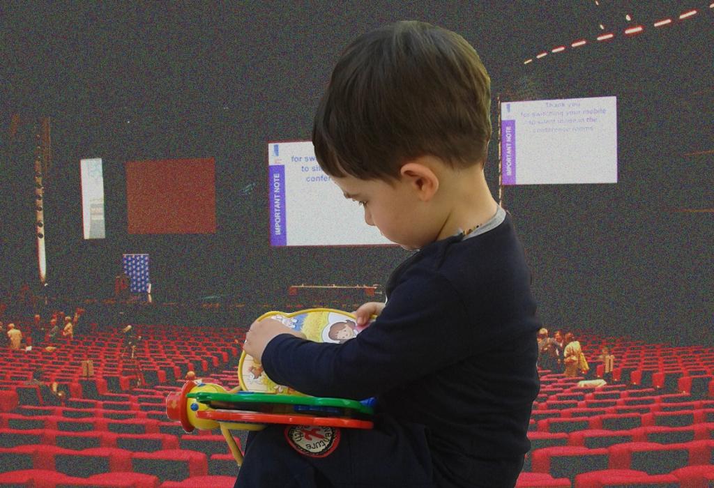 un bambino legge un librino, sullo sfondo una sala congressi con effetto sfocato e anticato